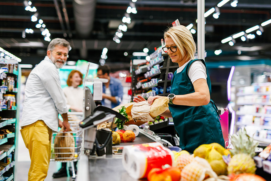 Une caissière senior scanne les courses d'un client à la caisse d'un supermarché, avec un homme senior poussant son caddie et attendant de payer ses courses.