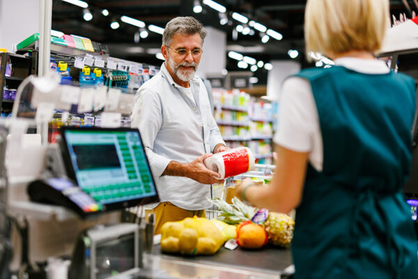 Senior man holding a pack of toilet paper, buying groceries at checkout counter in a supermarket, interacting with cashier