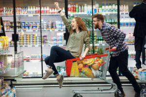 Happy couple having fun while choosing food in the supermarket. Young happy man pushing shopping cart with his girlfriend inside